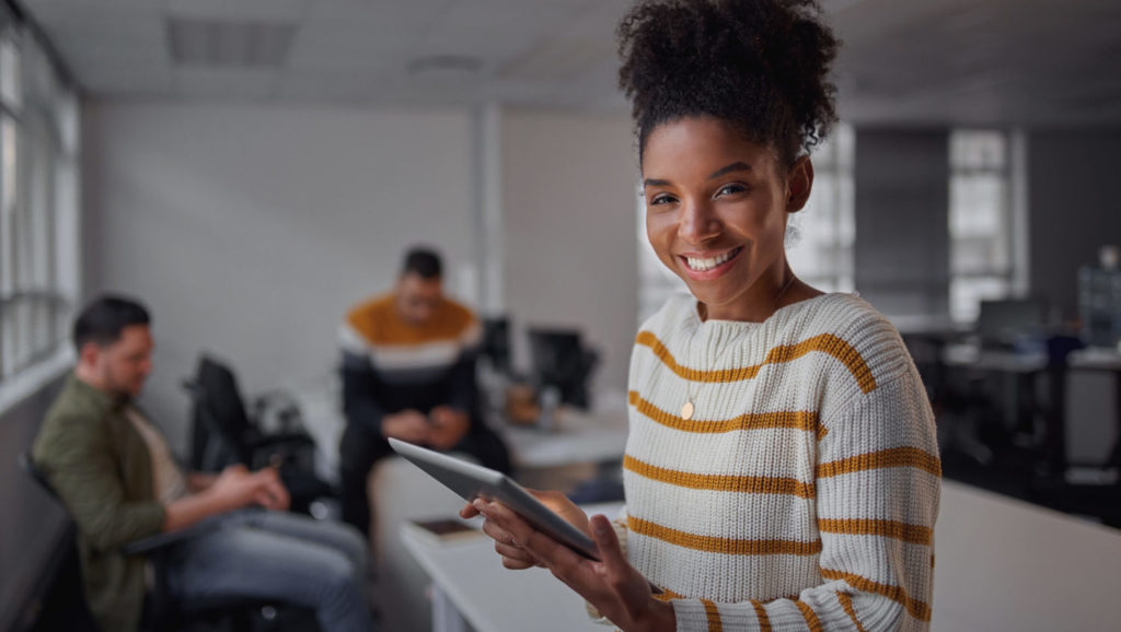 Women smiling holding tablet in an office setting with coworkers behind.