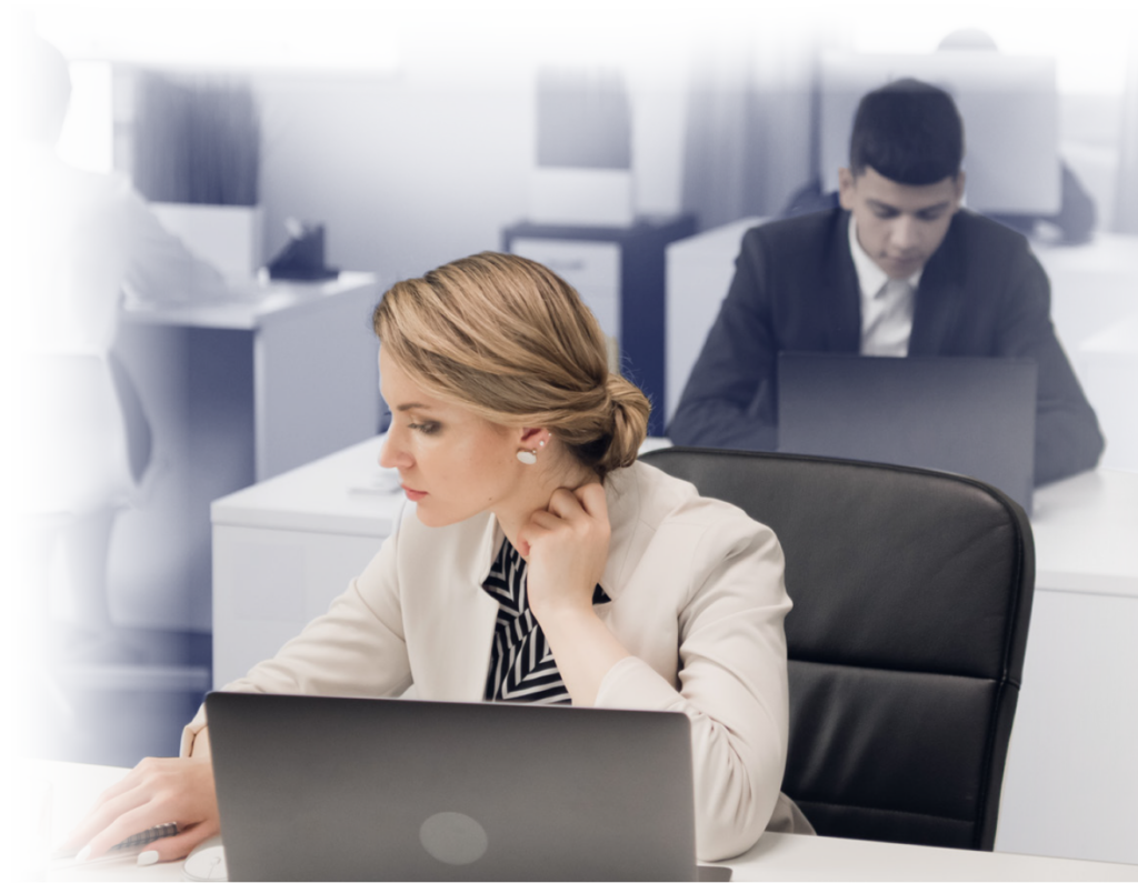 Blond office worker with hair in a bun looking at a paper beside her laptop with a man working at the desk behind her.